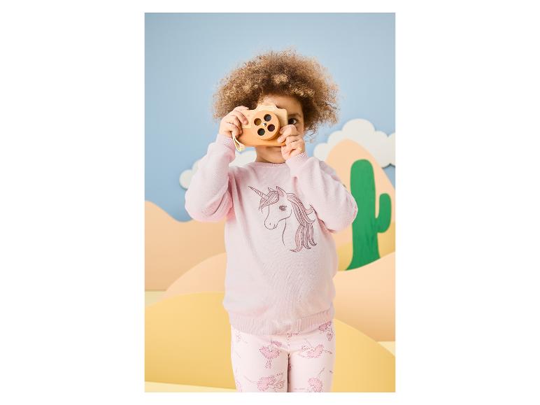 Fille aux cheveux bouclés en pull rose licorne et pantalon ballerine rose, jouant avec un appareil photo en bois.