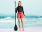 Une femme avec une pagaie de paddleboard, porte un legging noir et un short rouge de la marque Mistral.