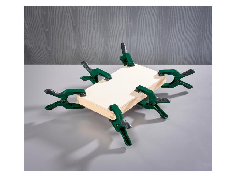 Six pinces à ressort vertes maintiennent un cadre en bois sur un fond gris.