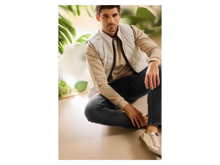 Homme en gilet matelassé beige, sweatshirt et jean foncé, assis dans un cadre naturel.