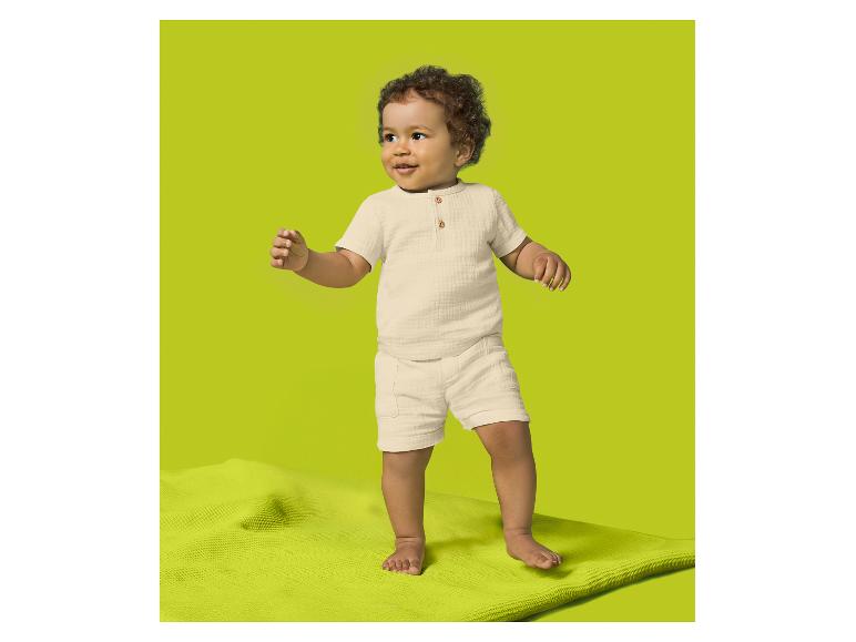 Bébé en ensemble chemise à manches courtes et short couleur crème, debout sur un tapis vert.