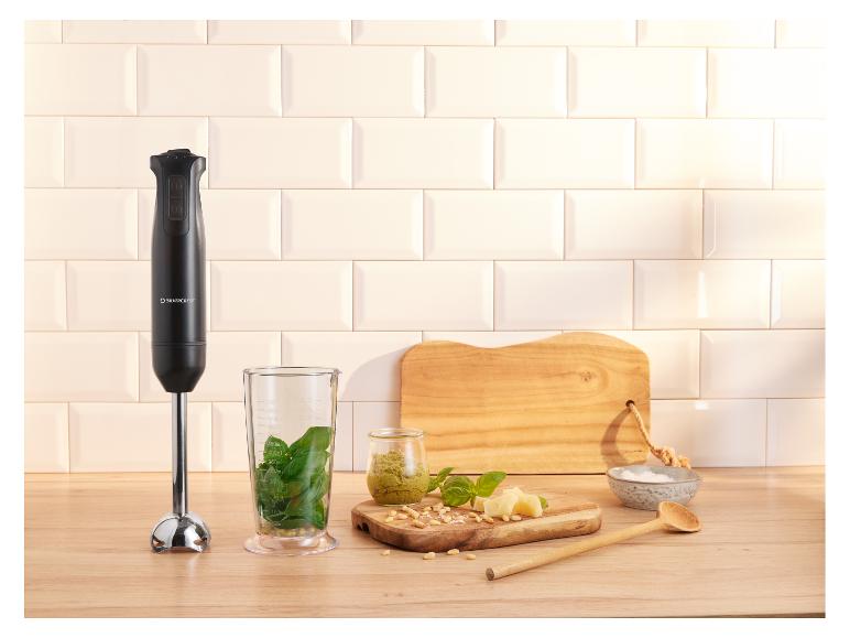 Mixeur plongeant avec ingrédients pour le pesto sur un plan de travail en bois.