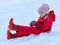 Un enfant en combinaison de neige rouge et bonnet rose assis sur une luge rouge dans la neige.