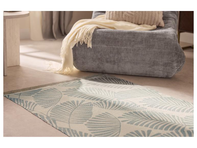 Fauteuil gris avec plaid et coussin beiges, tapis clair à motif de feuilles bleues sur le sol.