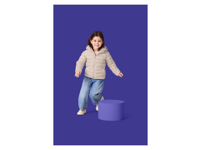 Fille souriante en doudoune beige et jean, sautant à côté d'un cylindre violet.