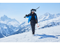 Homme en veste de ski, pantalon et casque, portant des skis et des bâtons sur la neige.