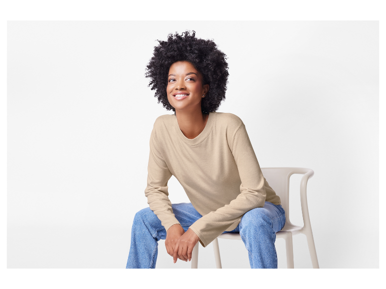 Femme souriante portant un pull beige et un jean, assise sur une chaise.