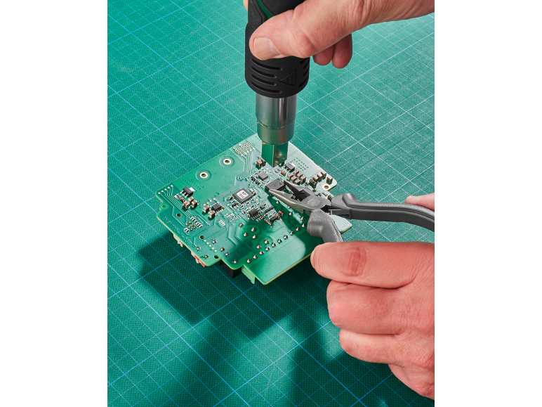 Mains réparant une carte de circuit imprimé avec un pistolet à air chaud et une pince sur un tapis de découpe.