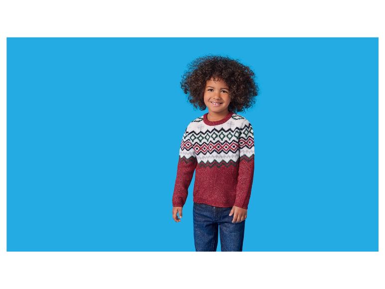 Un garçon souriant aux cheveux bouclés, portant un pull rouge et blanc à motifs et un jean.