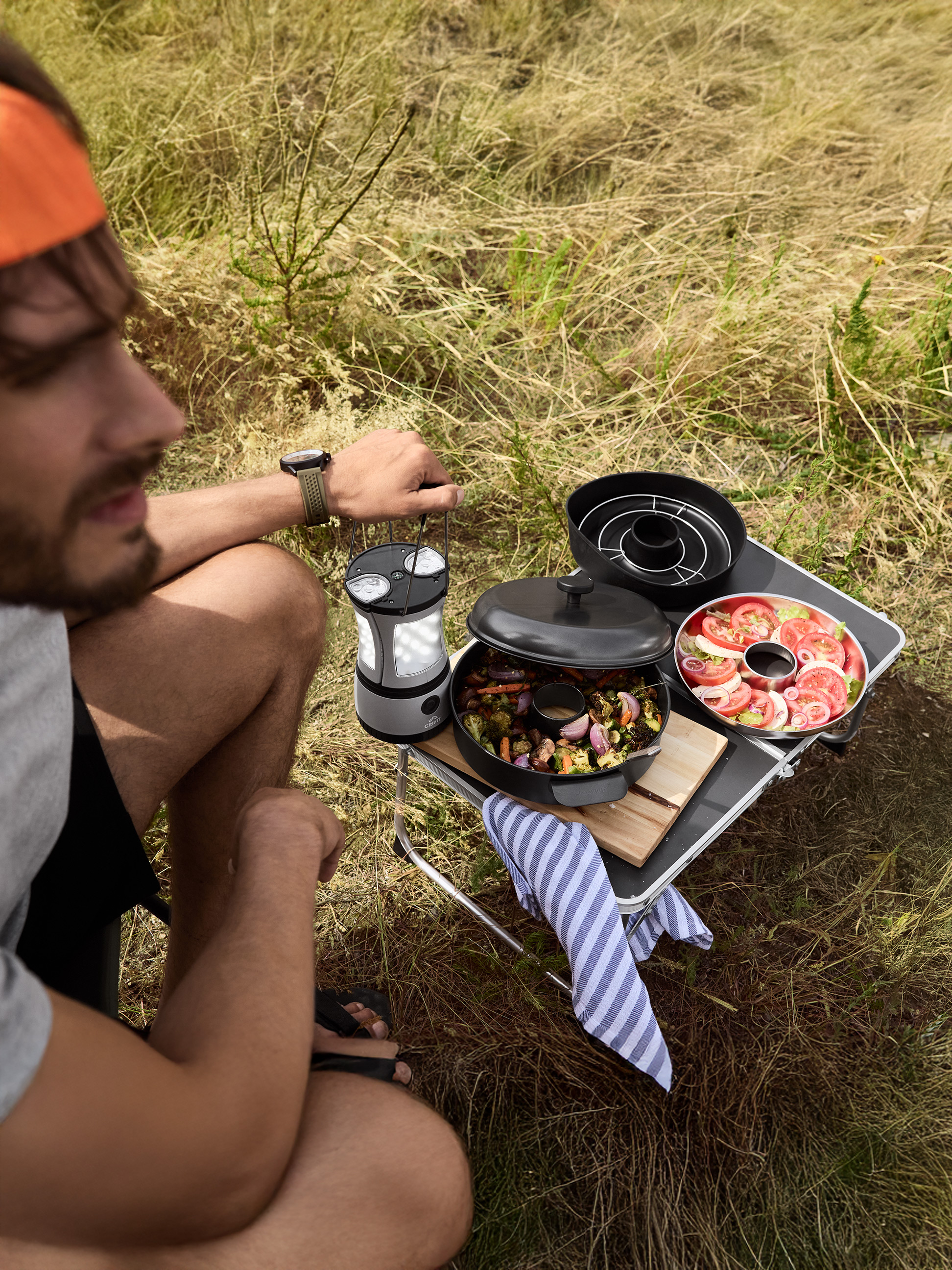 Homme cuisinant en extérieur avec un grill portable, servant des légumes rôtis et une salade de tomates.