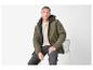 Homme en veste vert olive, pull beige et jean gris, assis sur une chaise.