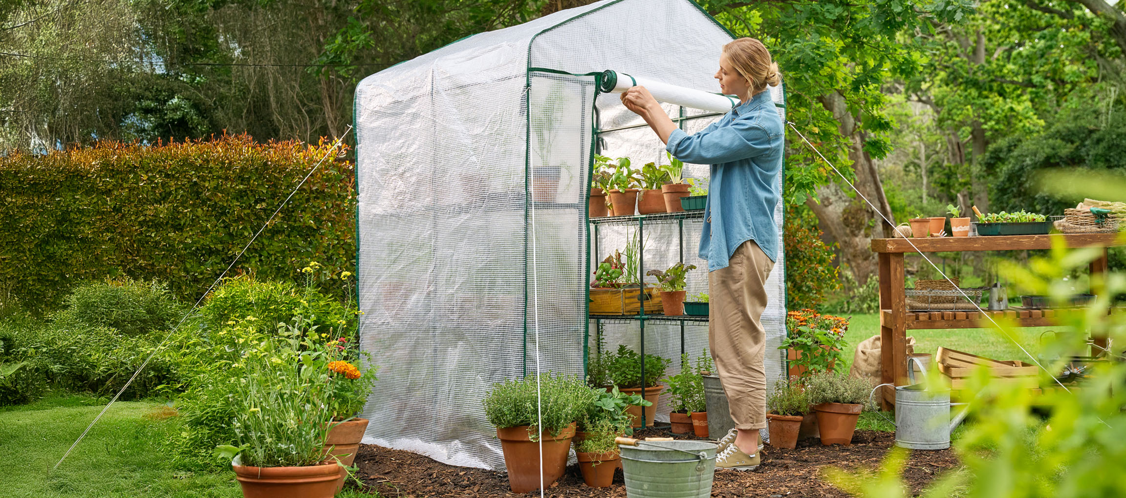Femme ouvrant une serre avec des plantes et des pots dans un jardin.