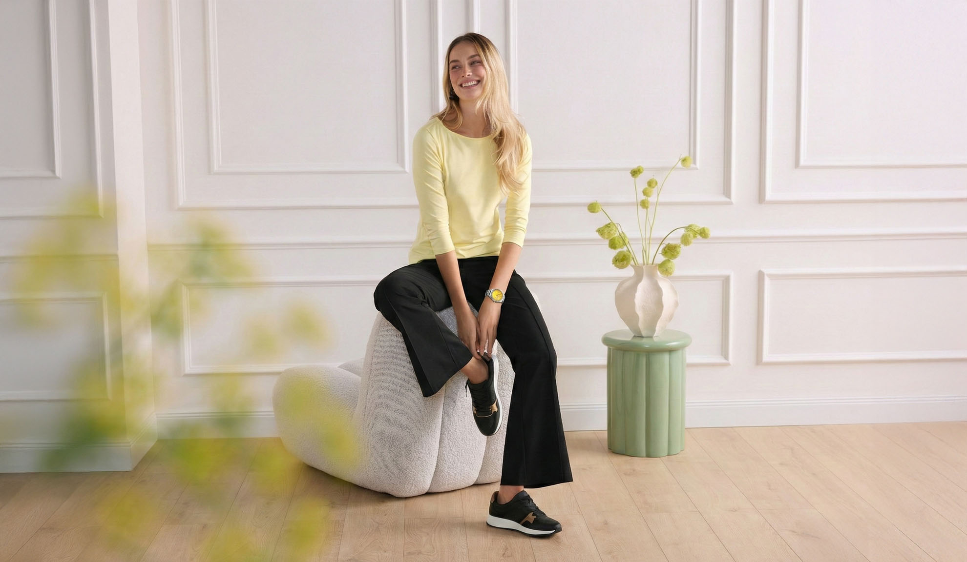Femme en haut jaune à manches longues et pantalon noir, assise sur un pouf, portant des baskets.