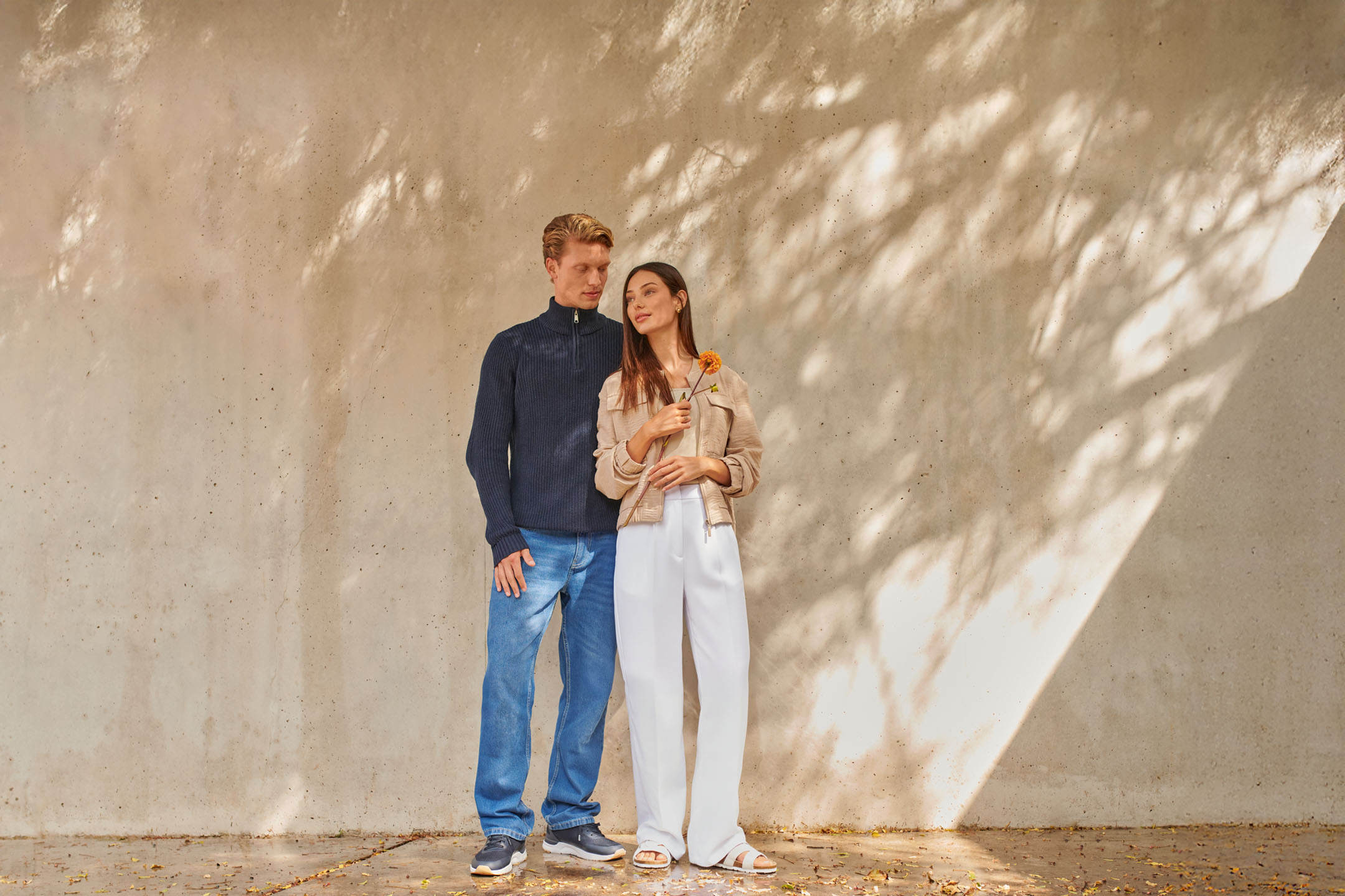 Couple en tenue décontractée: homme en pull bleu et jean, femme en veste beige et pantalon blanc.