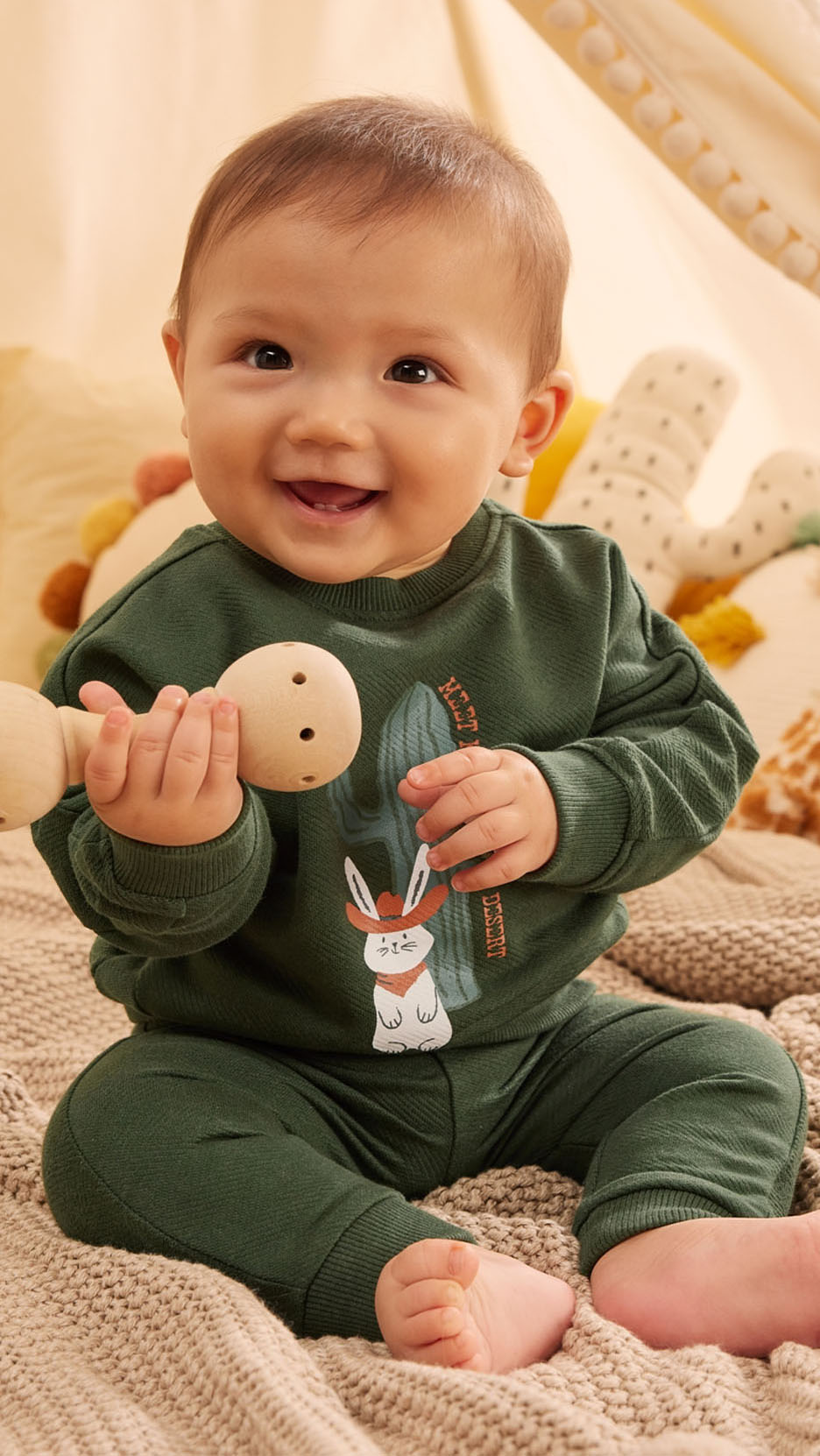 Un bébé souriant porte une tenue vert olive avec un imprimé de lapin western.