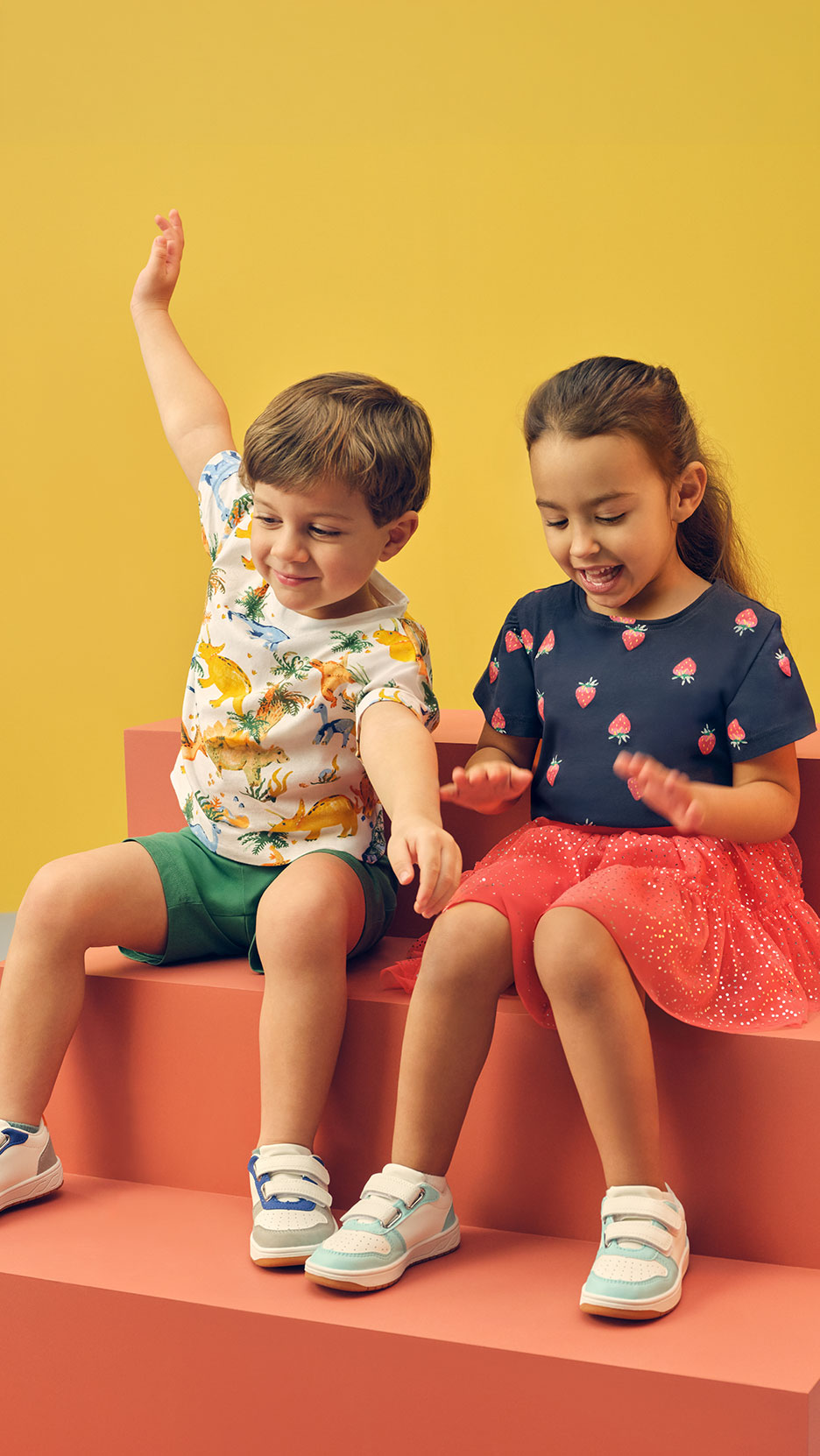 Deux enfants assis sur des marches, un garçon en t-shirt dinosaures, une fille en t-shirt fraises.
