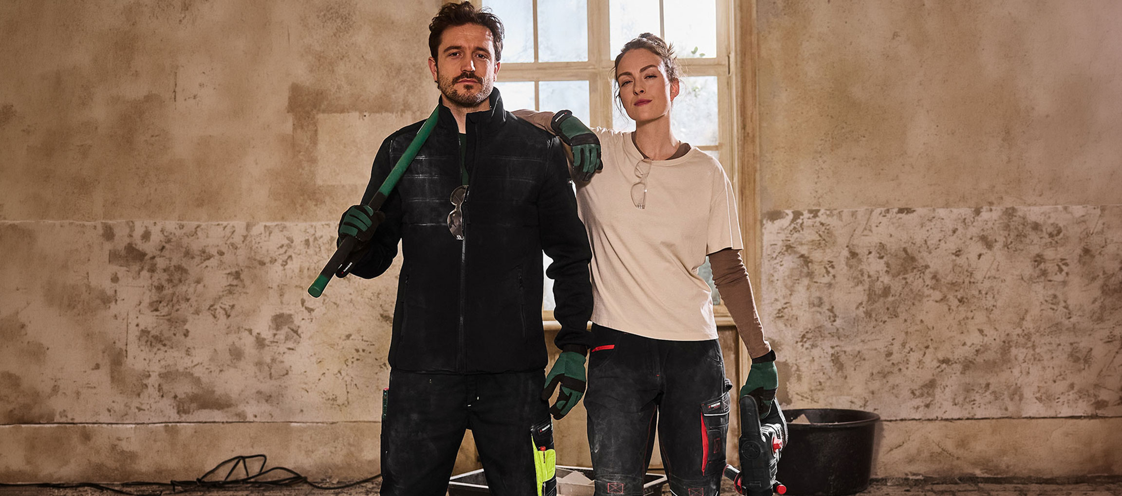 Homme et femme en tenue de travail, outils en main, dans une pièce en rénovation.