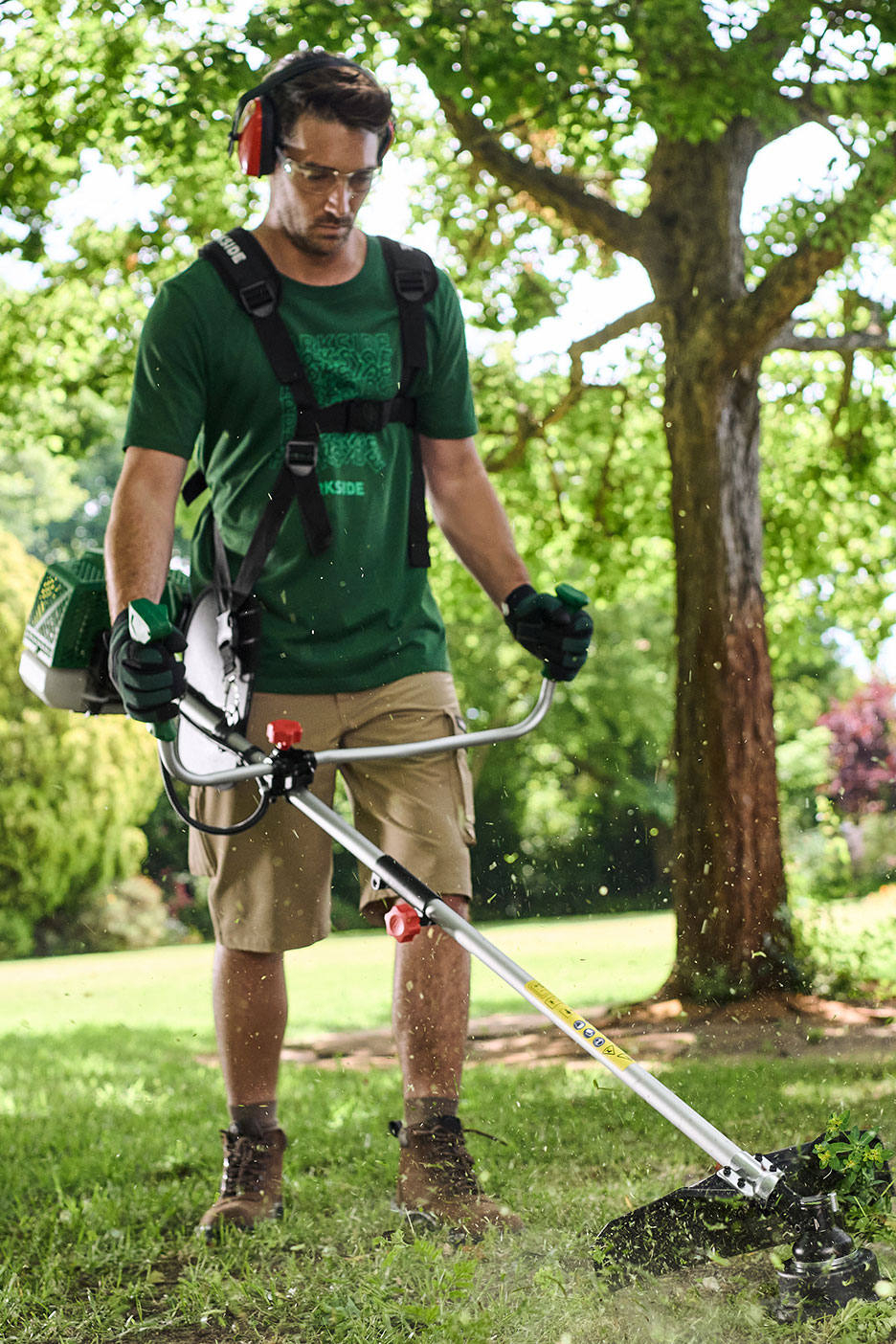Homme utilisant une débroussailleuse avec harnais, casque anti-bruit et lunettes de protection dans un jardin.