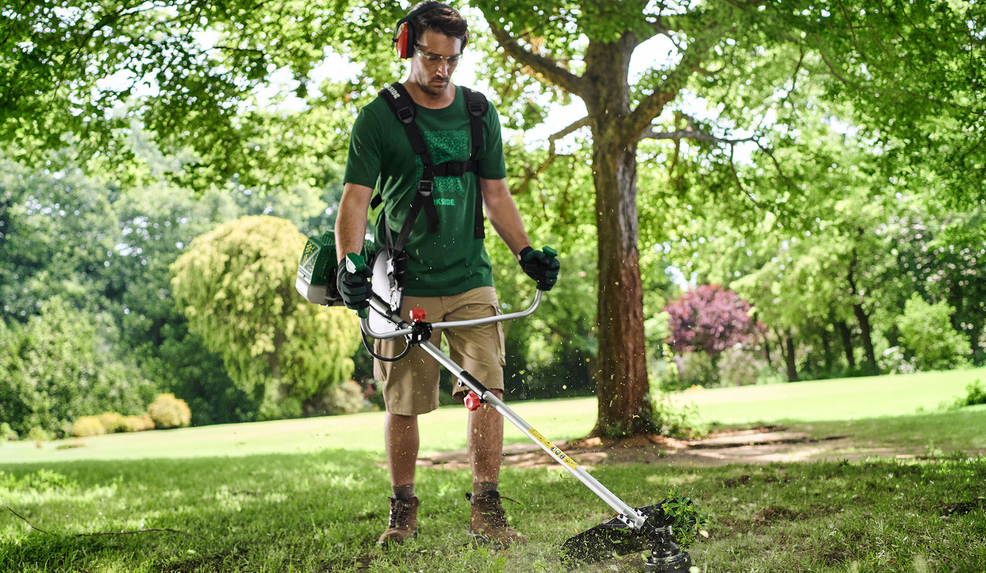 Homme coupant l'herbe dans un jardin avec une débroussailleuse, portant lunettes et casque.