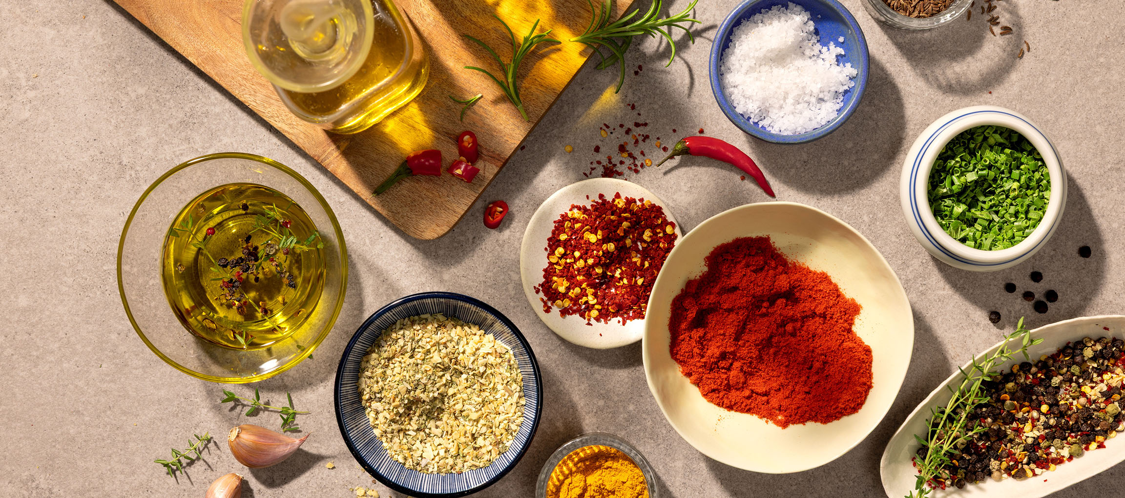 Variété d'épices et d'huiles, dont paprika, piment, sel, poivre et huile d'olive, sur fond gris.