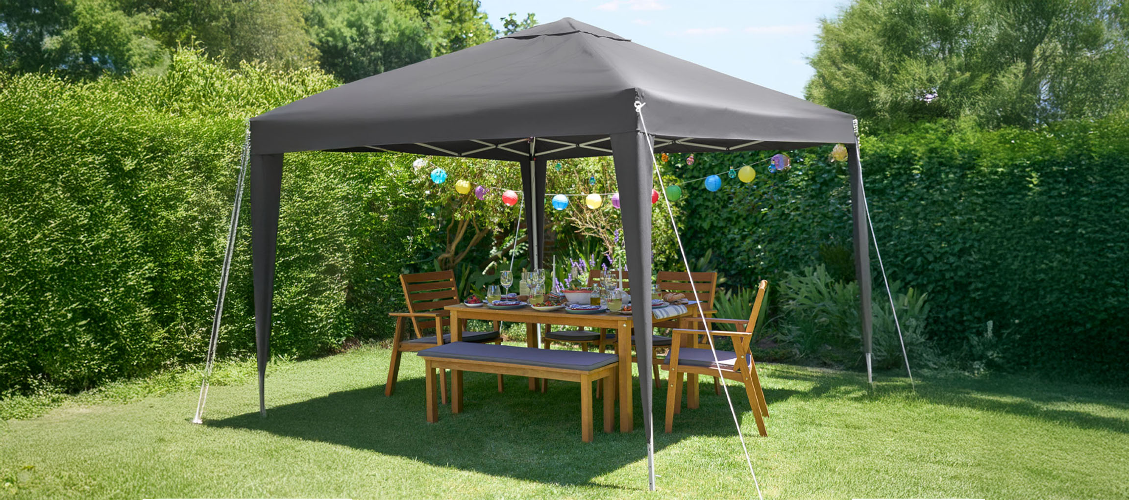 Un gazebo gris foncé escamotable abrite un ensemble de salle à manger en bois d'extérieur dans un jardin verdoyant luxuriant.