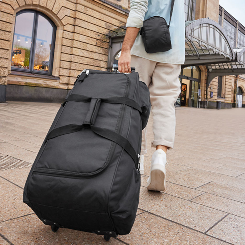 Un voyageur tire une grande valise noire à roulettes devant une gare.