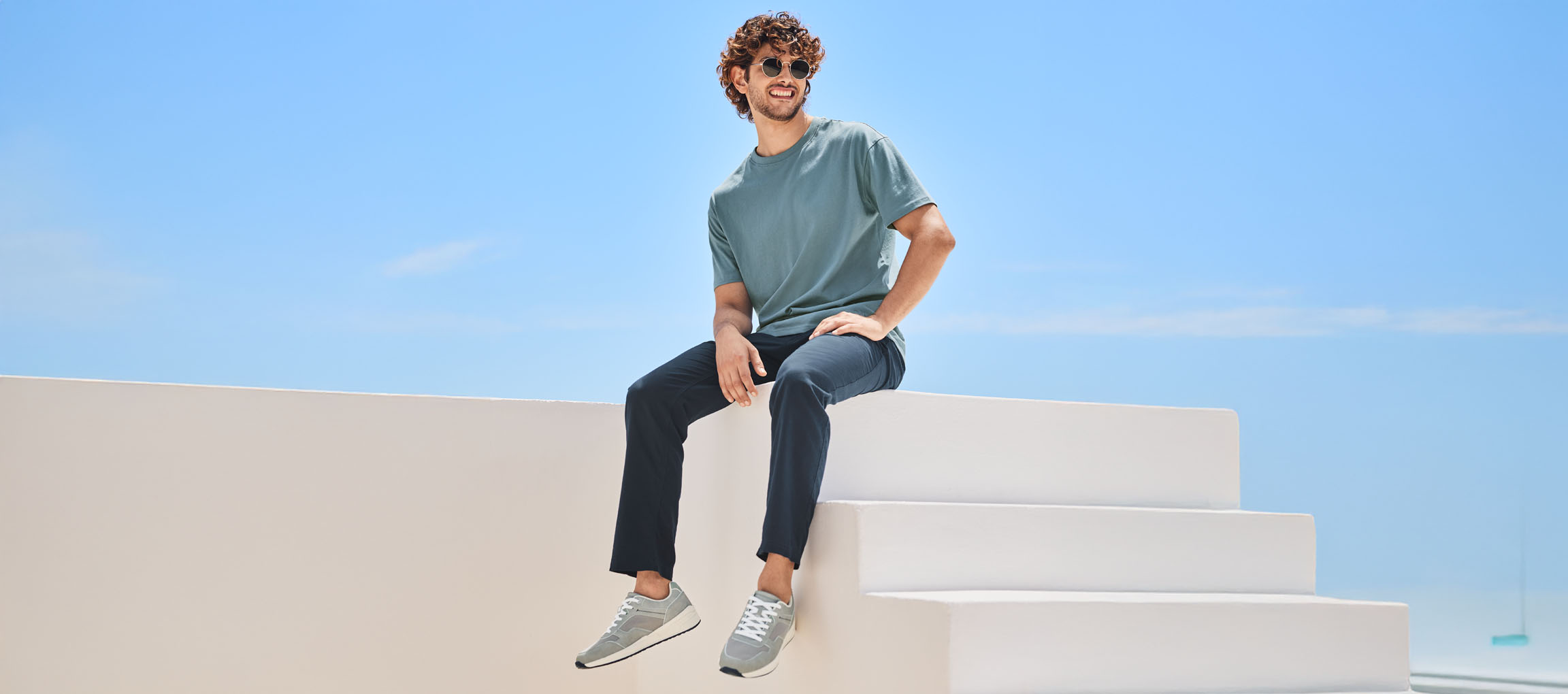 Homme détendu portant un t-shirt, un pantalon et des New Balance, assis sur un escalier blanc.