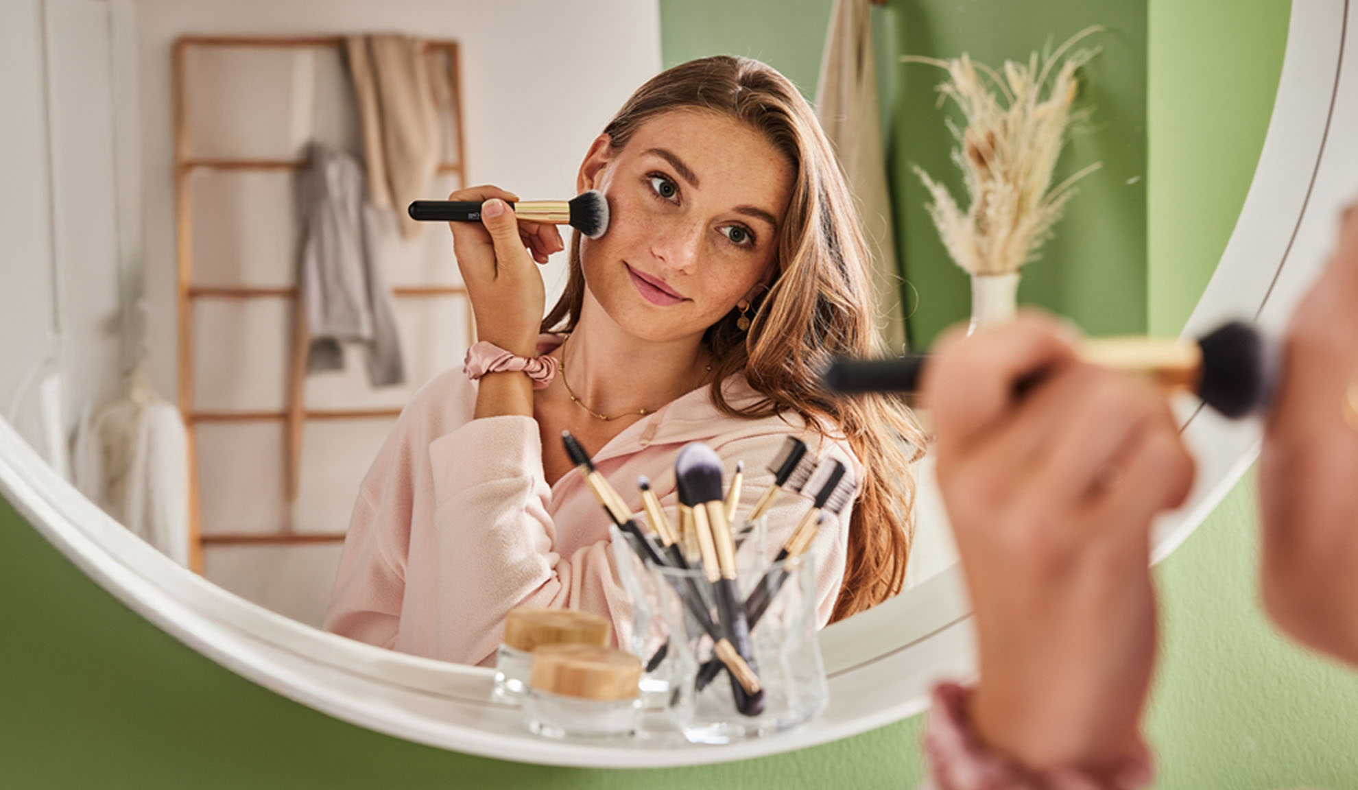 Femme appliquant du maquillage avec un pinceau, reflétée dans un miroir ovale.