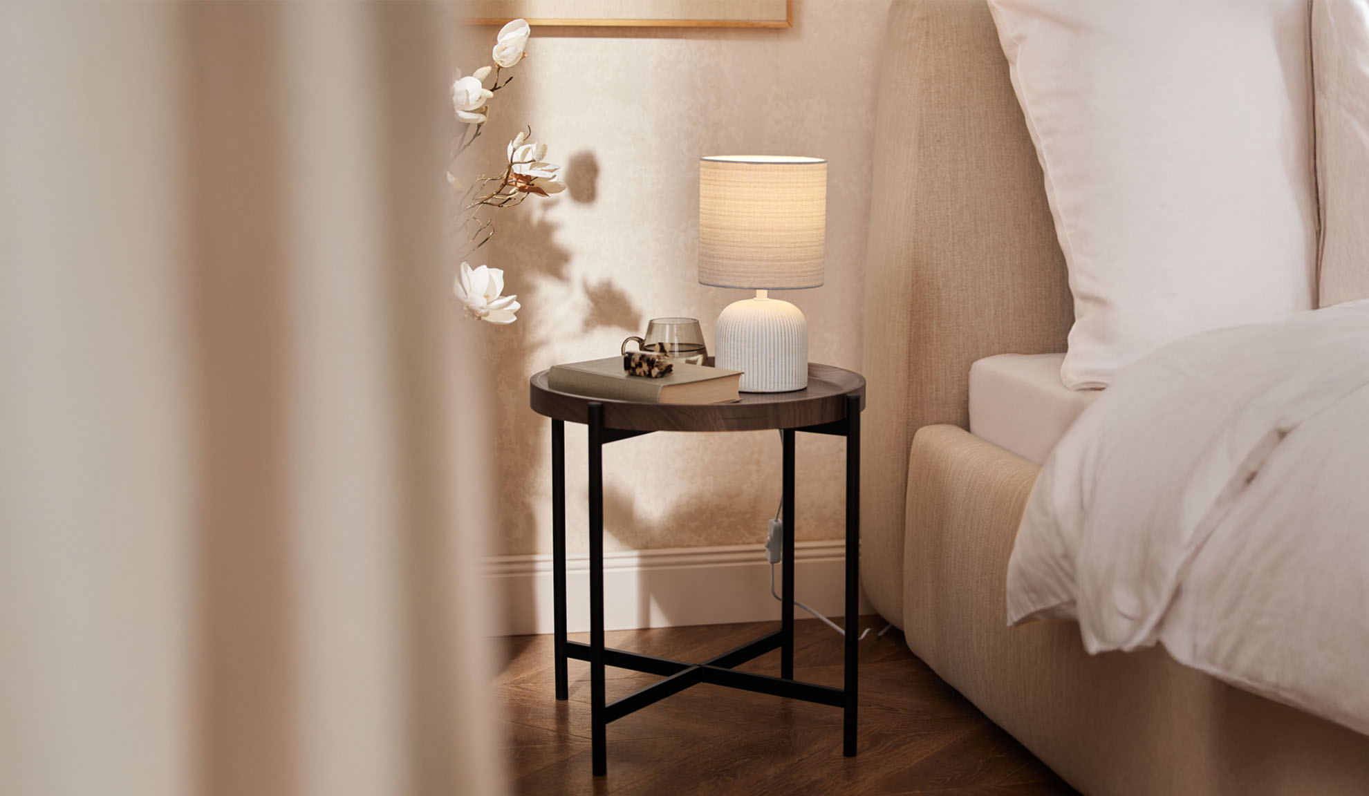 Table de chevet ronde en bois avec lampe de table et accessoires près d'un lit.
