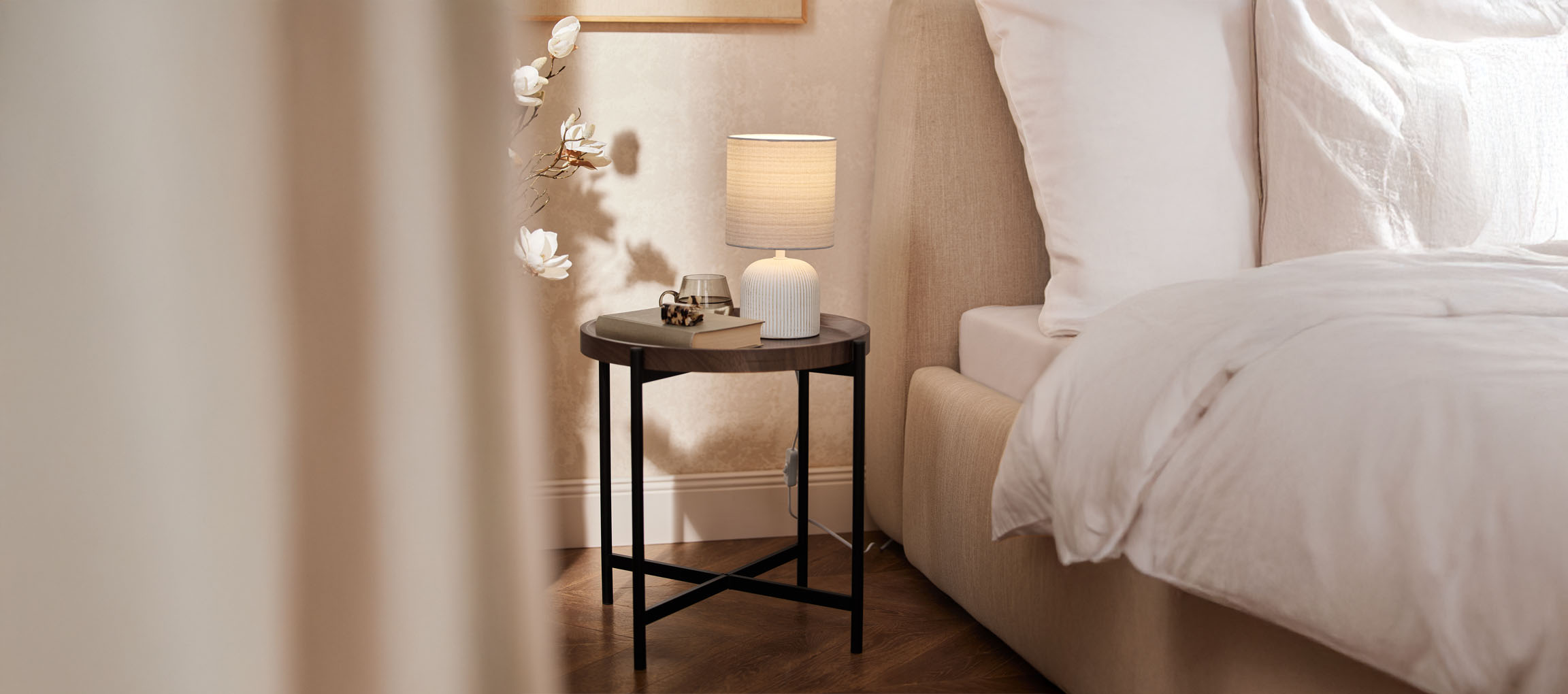 Table de chevet ronde en bois avec lampe de table et lit beige dans une chambre douillette.
