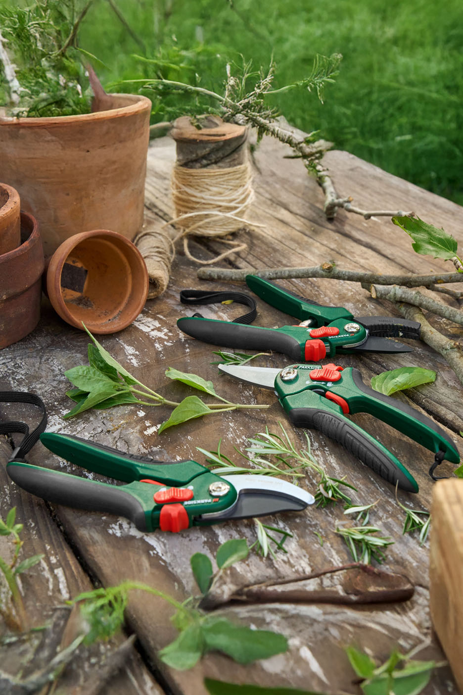 Trois sécateurs Parkside sur une table en bois avec des pots et des herbes.