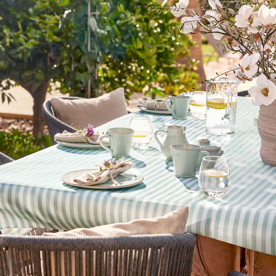 Table dressée avec vaisselle et verres sur une nappe rayée verte en terrasse.