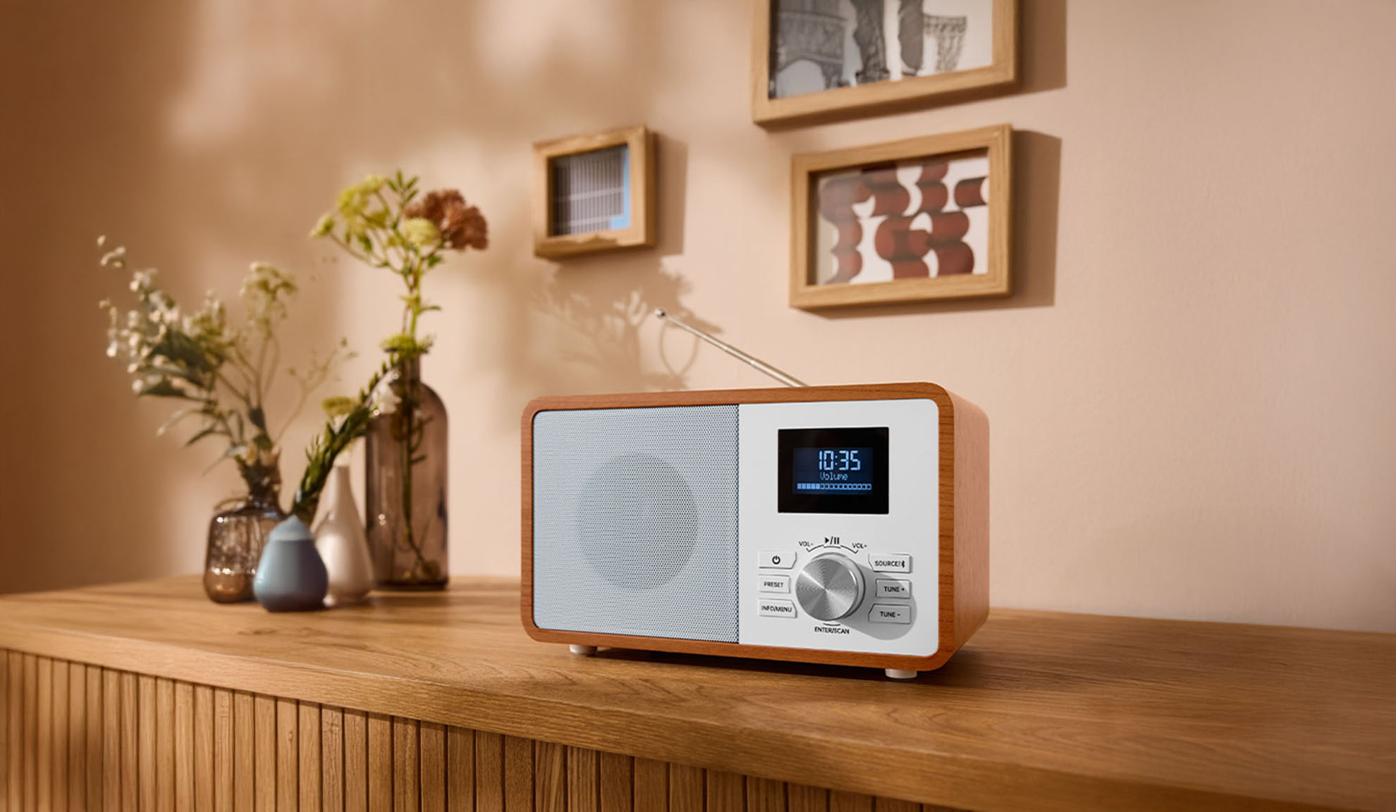 Une radio numérique en bois sur une commode, avec des vases et des cadres photo en arrière-plan.