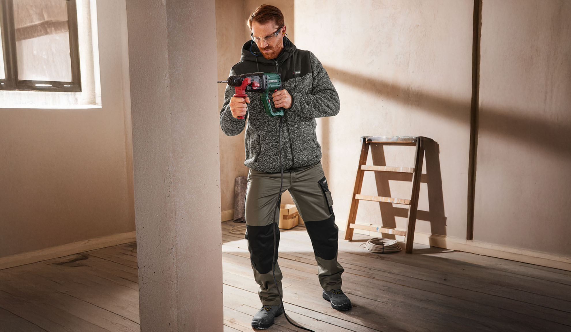 Homme en veste et pantalon de travail, tenant une perceuse dans une pièce inachevée.