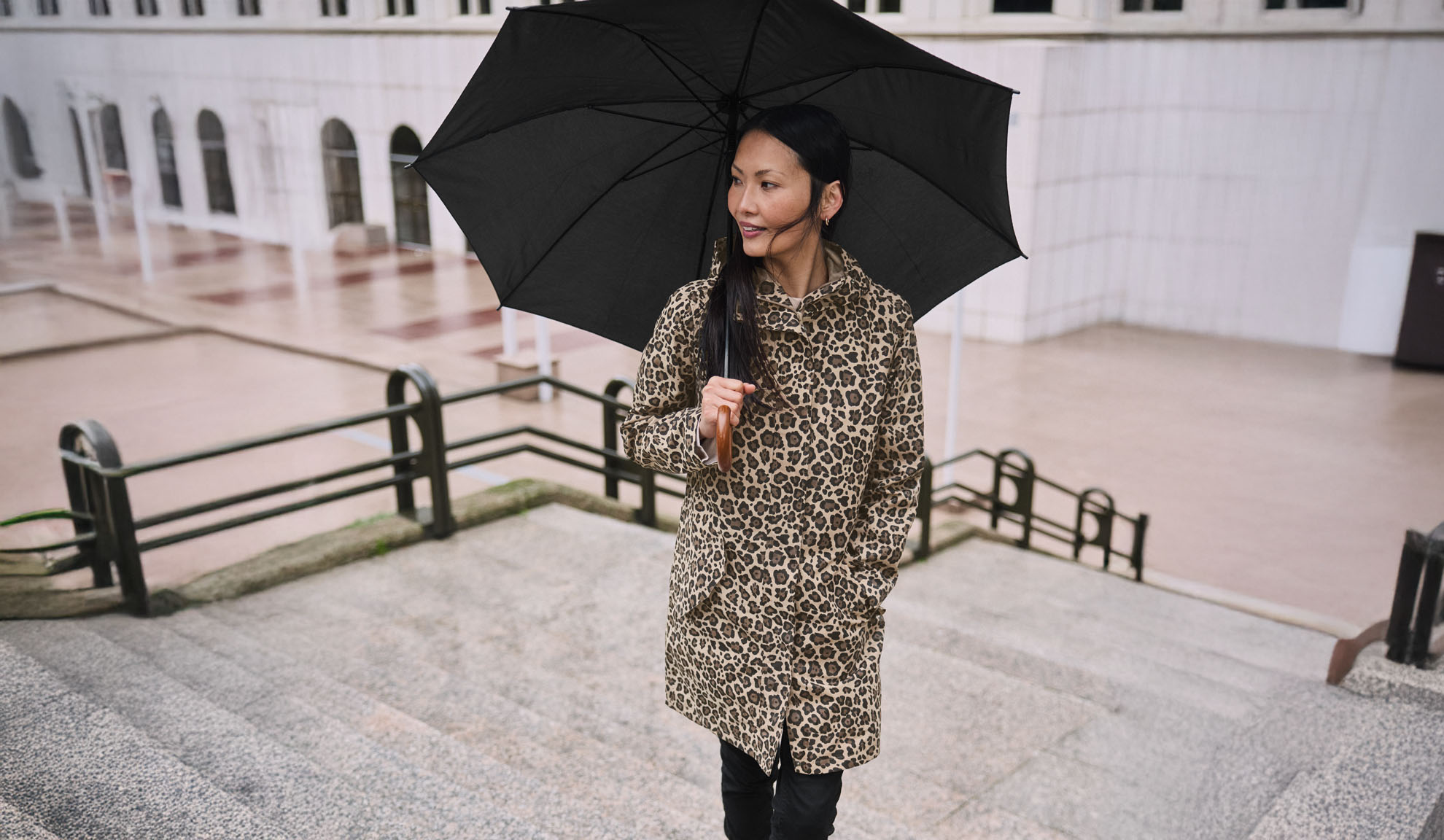 Une femme en manteau léopard tient un parapluie noir dans un escalier.