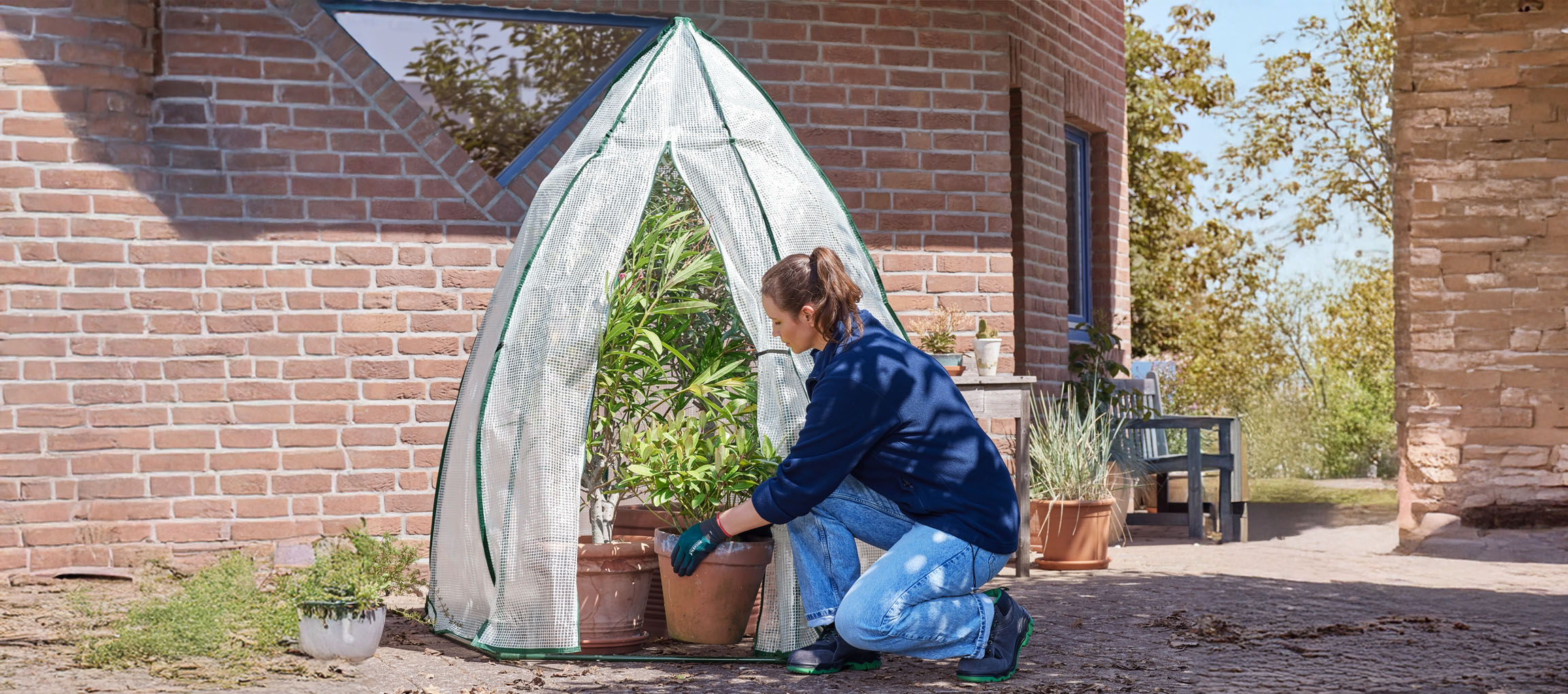 Femme plaçant des plantes en pot dans une serre près d'un bâtiment en briques.