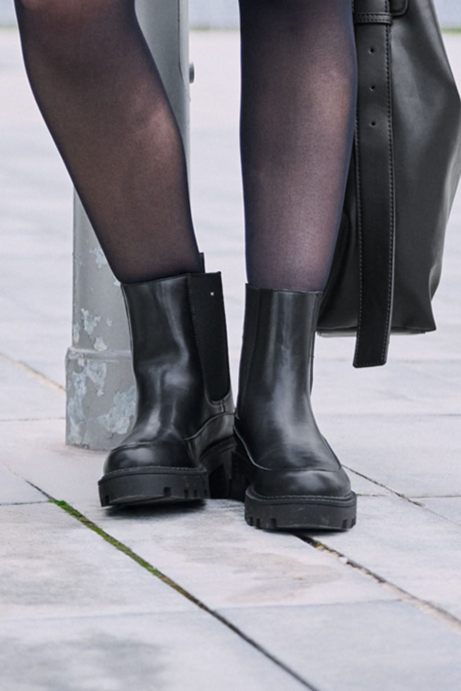 Jambes de femme en collants noirs et bottes Chelsea noires, avec un sac noir.