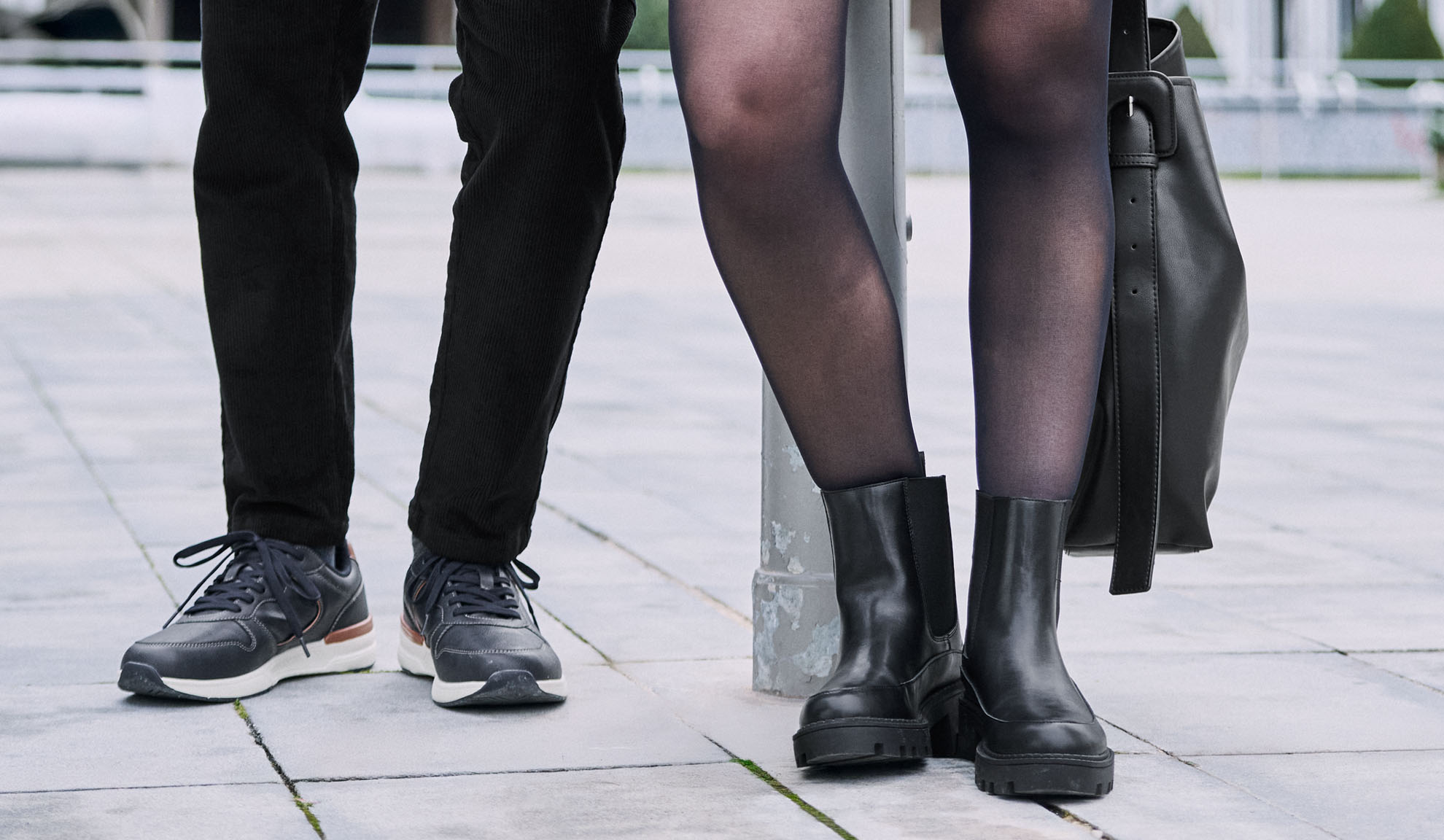 Jambes de deux personnes, l'une portant un pantalon et des baskets, l'autre des collants, des bottes et un sac.