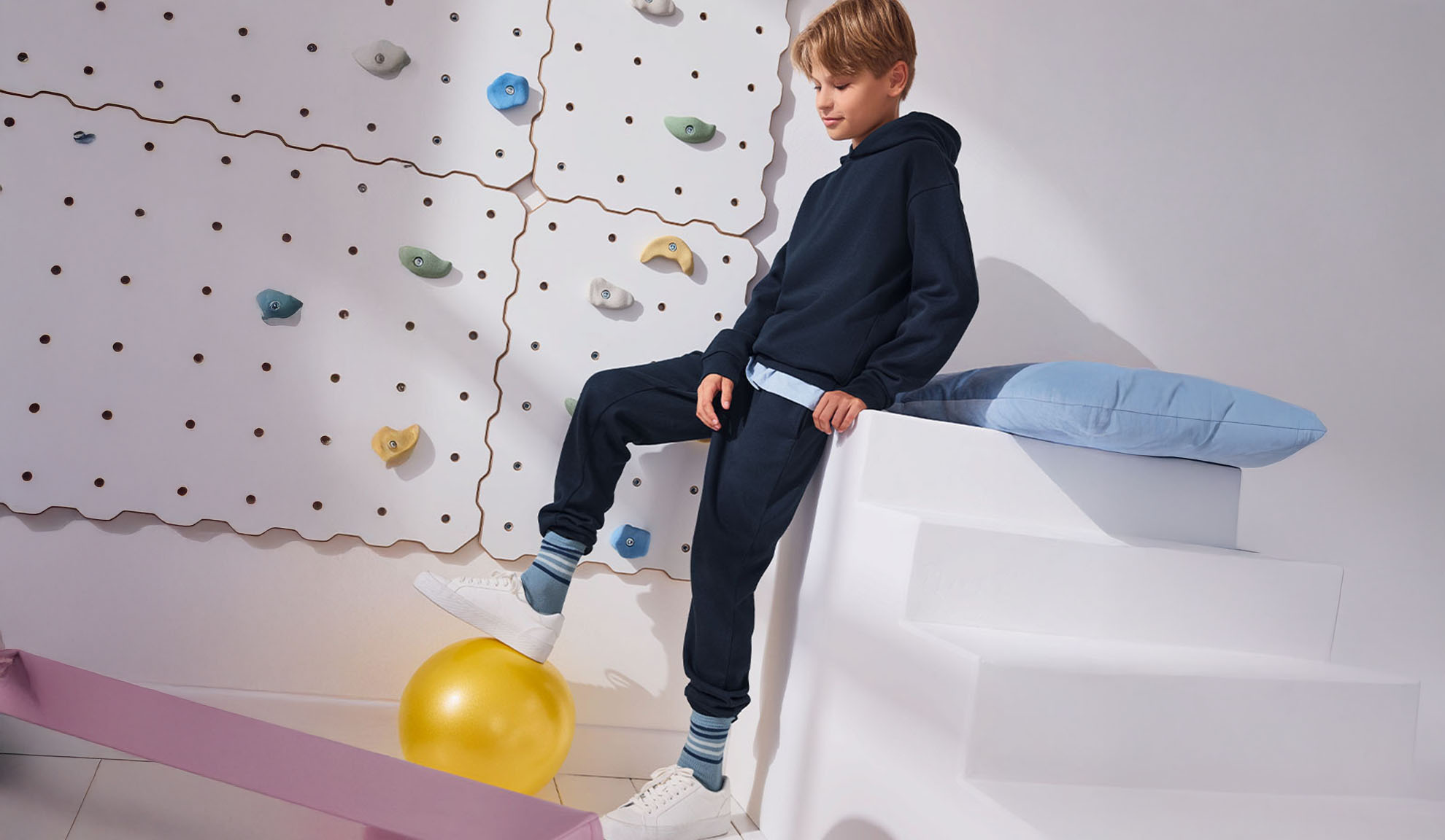 Un garçon debout sur un ballon d'exercice près d'un mur d'escalade, portant un sweat à capuche et un pantalon de survêtement.