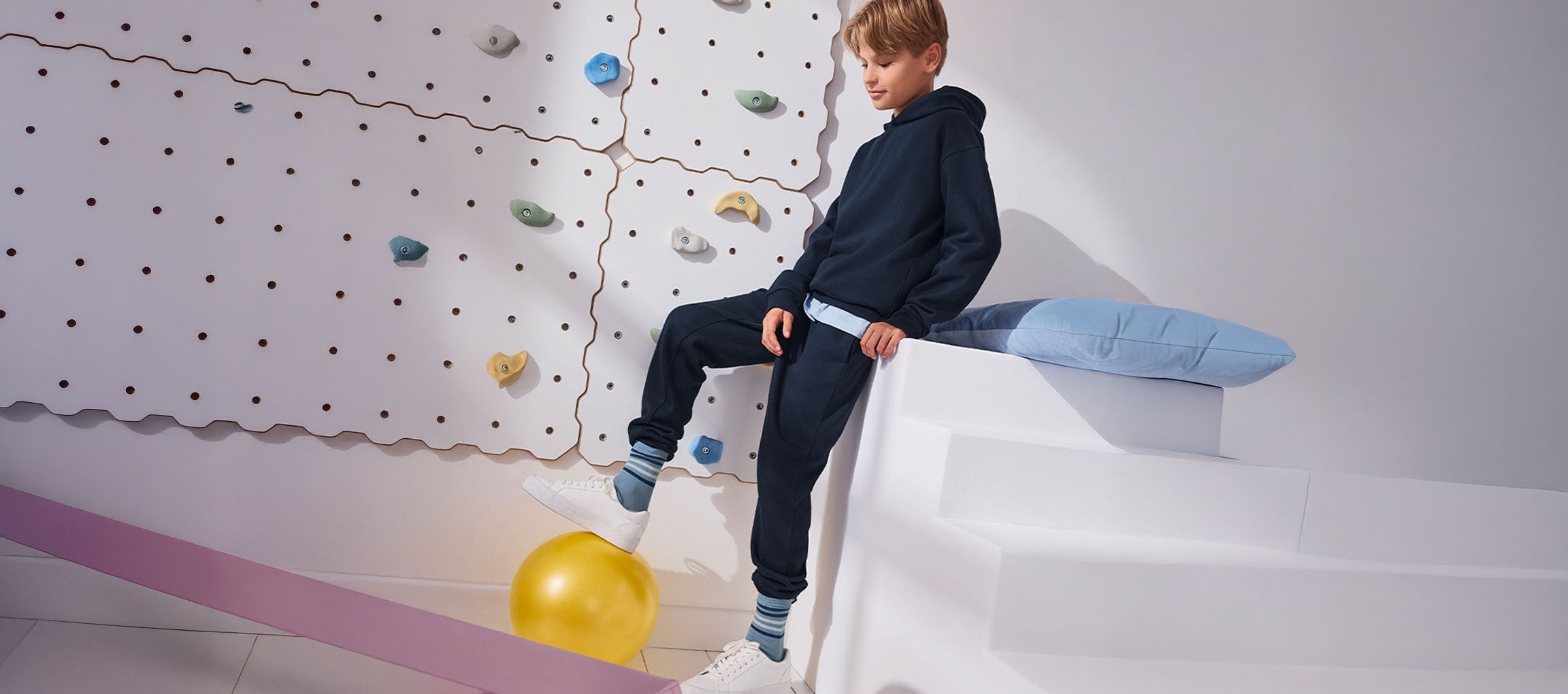Un garçon en sweat à capuche et pantalon de survêtement bleu marine, un pied sur un ballon jaune, à côté d'un mur d'escalade.