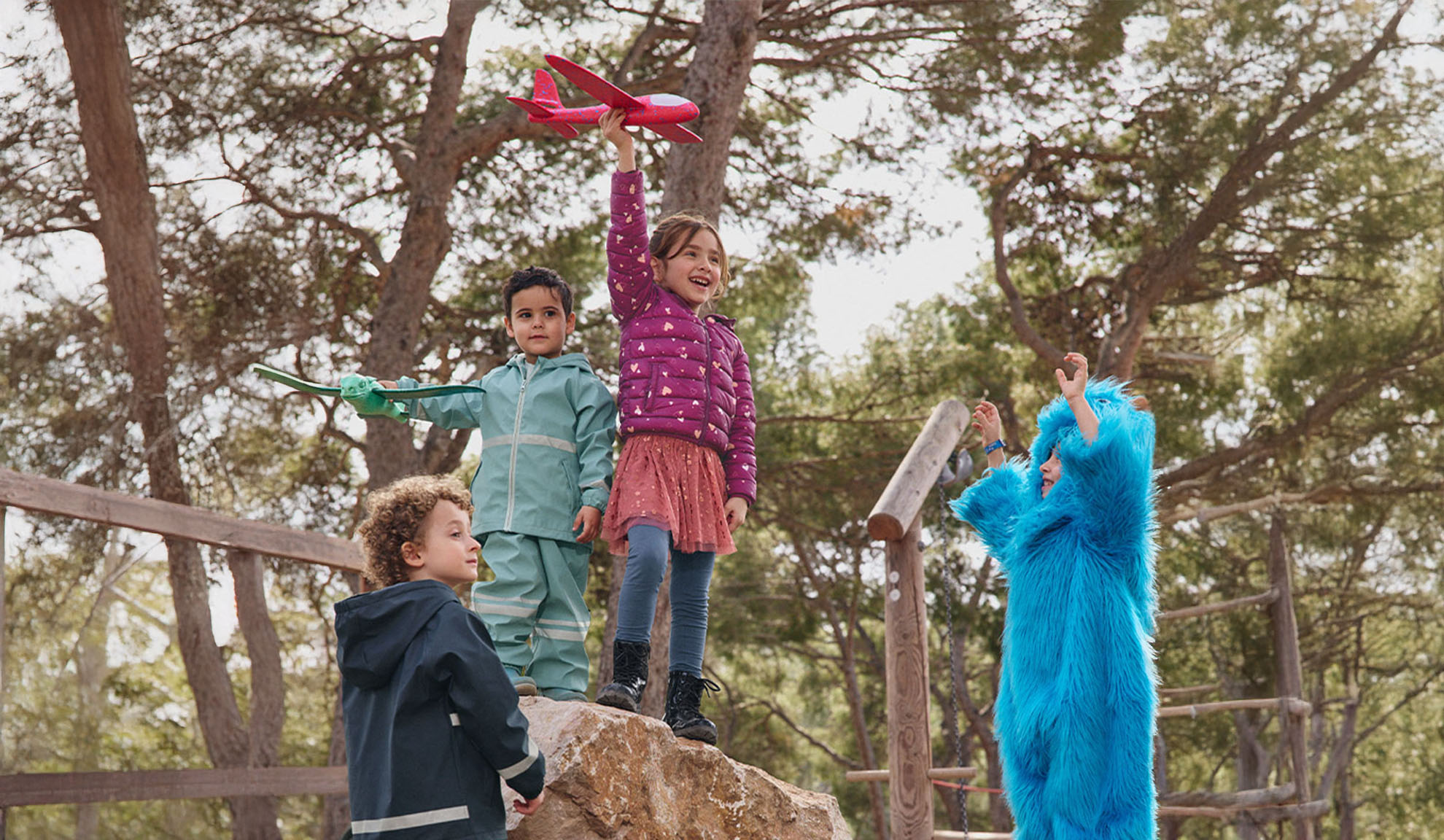 Quatre enfants jouent dehors, deux avec des avions jouets et un dans un costume de monstre bleu.