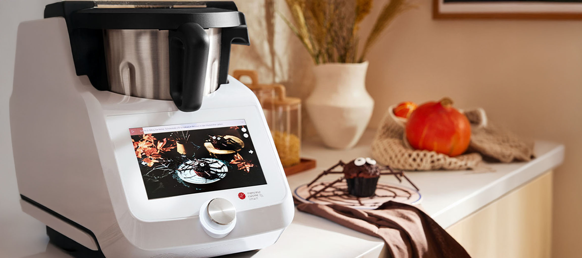 Robot de cuisine avec écran tactile affichant une recette d'Halloween, à côté d'un cupcake araignée.