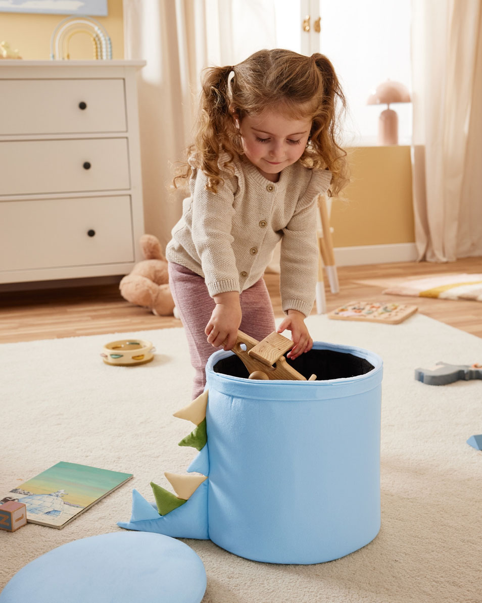 Une enfant met des jouets en bois dans un bac de rangement bleu sur le thème des dinosaures.