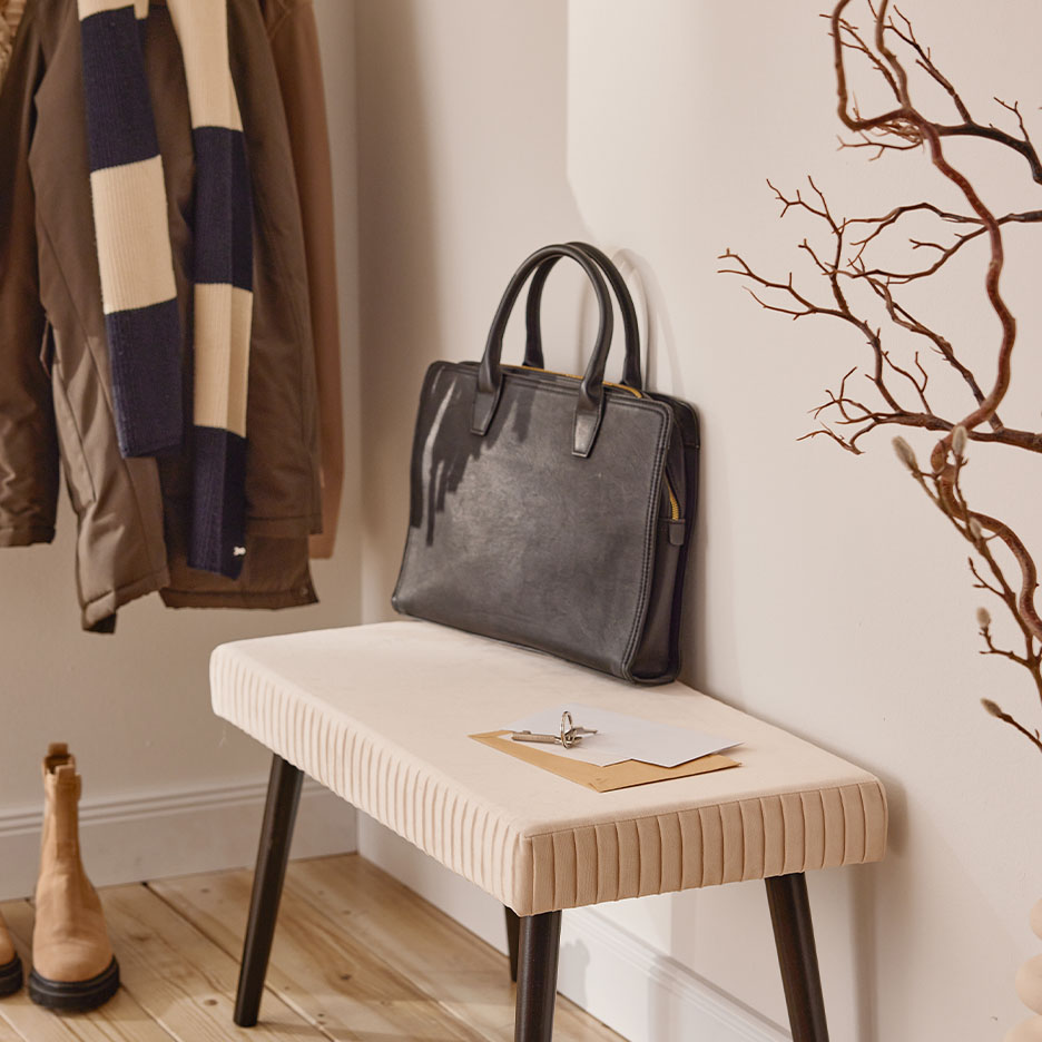 Un sac à main noir sur un banc beige dans un couloir avec un manteau et une écharpe.
