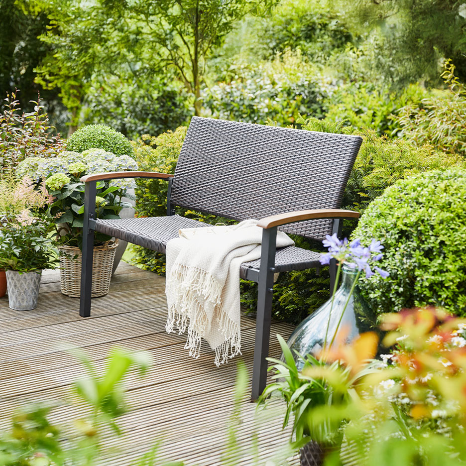Un banc d'extérieur gris foncé avec une couverture sur une terrasse en bois entourée de plantes luxuriantes.