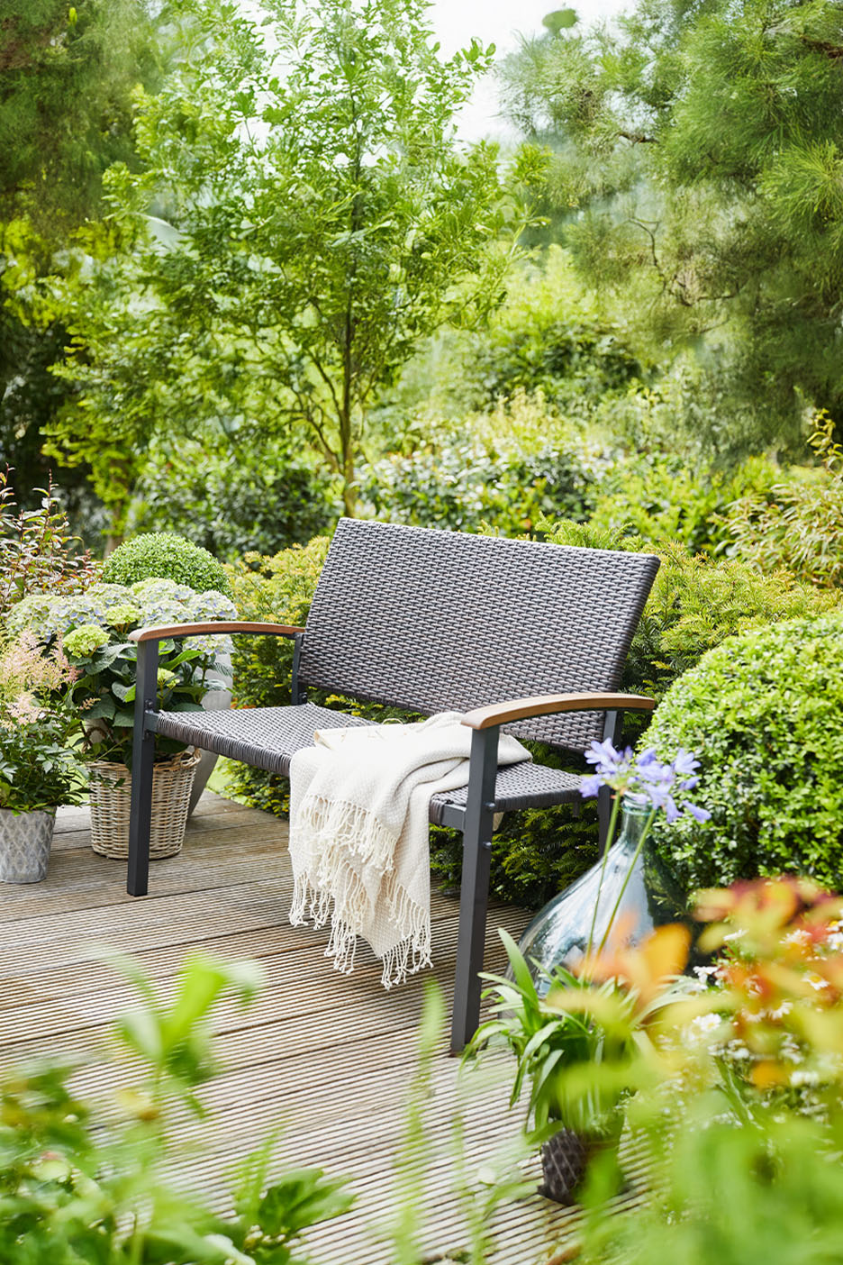 Banc de jardin en rotin avec une couverture sur une terrasse en bois, entouré de plantes.