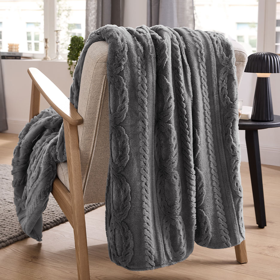Plaid gris en tricot torsadé drapé sur un fauteuil beige dans une pièce douillette.