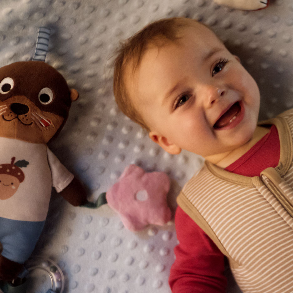 Bébé souriant allongé à côté d'une peluche loutre et d'un jouet rose en forme de fleur.