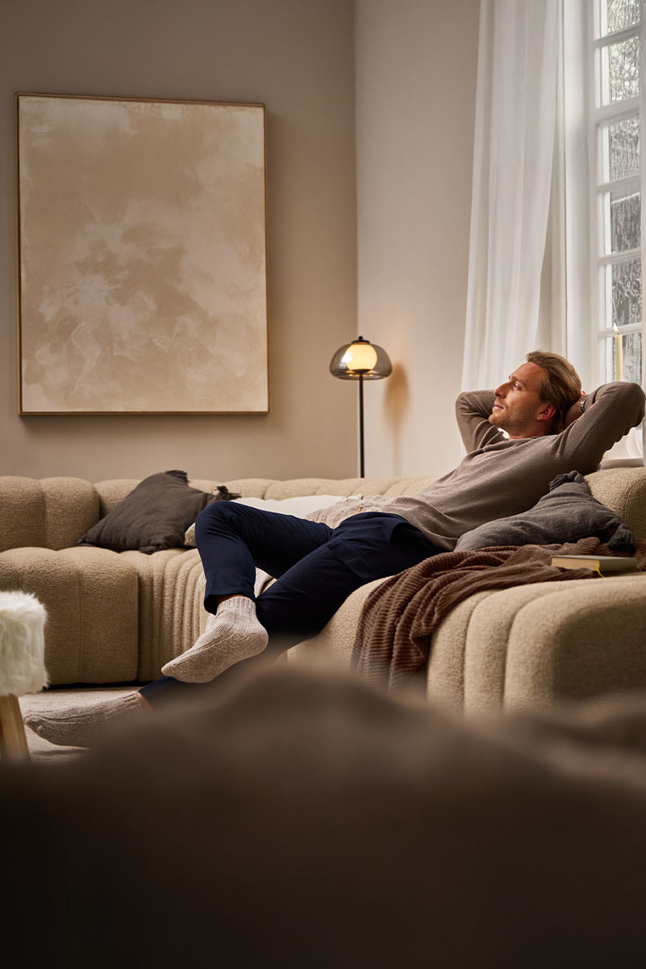 Un homme se détend sur un canapé beige dans un salon confortable avec une peinture abstraite et un lampadaire.