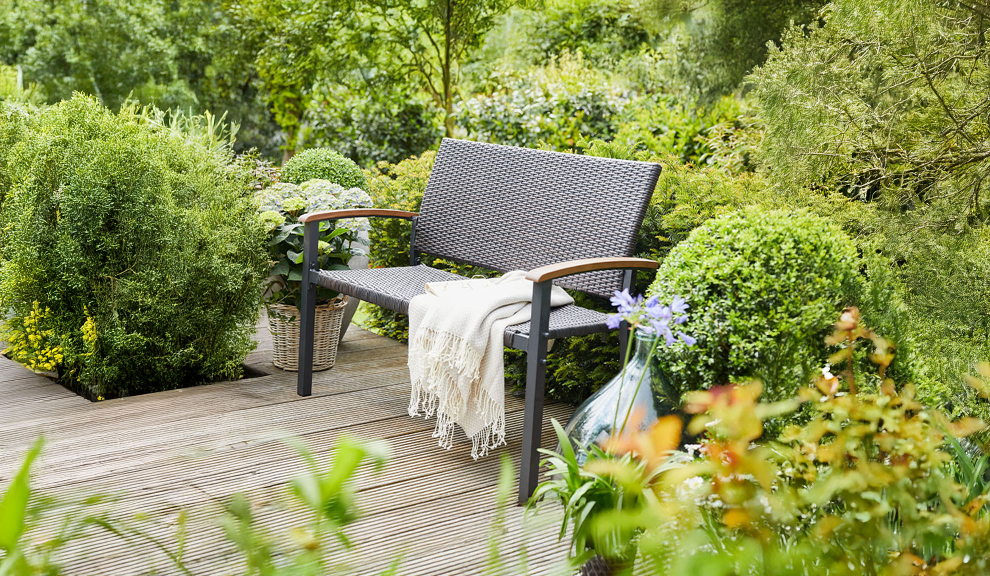 Banc de jardin en rotin sur une terrasse en bois, entouré de verdure luxuriante.